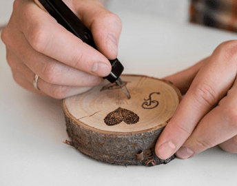 A close up image of a pair of hands burning a heart and other shapes into a wood coin using a wood burning tool